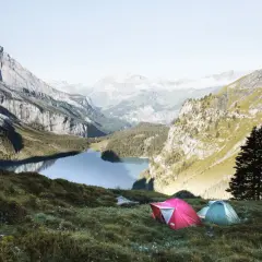 Two tents are set up on a grassy slope overlooking a serene lake in the Swiss Alps. This is a perfect spot for swiss alps hiking and camping.