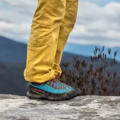 A person stands on a rock, wearing yellow pants and blue approach shoes with orange laces, ready for an adventure.