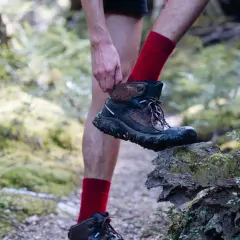 A hiker adjusts their muddy Keen shoes, showing signs of wear and tear, as they step onto a moss-covered log in a forest.