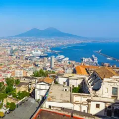One day in Naples, a panoramic view reveals the city's sprawl, the bustling port with cruise ships, and the majestic Mount Vesuvius in the distance under a clear blue sky.