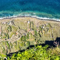 Terraced fields and houses dot the coastline, a glimpse of what to see in Madeira. The turquoise ocean crashes against the rocky shore.