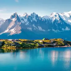 A serene lake reflects snow-capped peaks during a Mont Blanc trek. A tent sits on a grassy knoll, hinting at an overnight adventure.