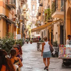 A woman walks down a bustling street in Sicily, past shops and cafes. The scene suggests a vibrant, lively atmosphere, answering the question: is Sicily safe for tourists to enjoy?
