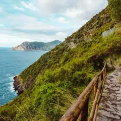 A stone path winds along the coast for Cinque Terre hiking, with lush green hills and the blue sea stretching to the horizon.