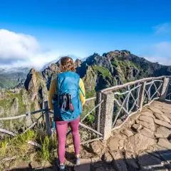 A hiker with a blue backpack enjoys a breathtaking view during a Madeira hike. Jagged, green mountains rise under a bright blue sky.