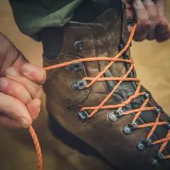 Close-up of hands tying orange laces through metal hooks on a brown hiking boot, demonstrating how to tie boots with hooks for a secure fit.