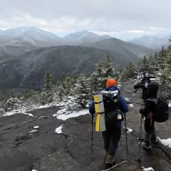 Hikers trek across a snowy mountain ridge, leaving no hiker trash behind. The vast, misty mountain range stretches into the distance under a cloudy sky.