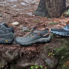 Three pairs of shoes sit on a mossy, wet forest floor: a mid-height hiking boot, a low-cut hiking shoe, and a pair of trail running shoes.