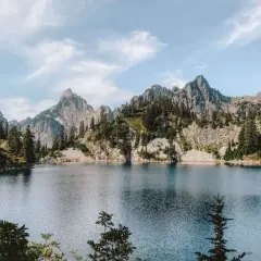 A serene alpine lake reflects jagged, tree-covered mountains. This view makes you wonder how long to hike 16 miles to reach such a peaceful spot.