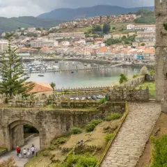 View from a castle wall overlooking a harbor filled with boats, a historic ship, and a town. The path leads through an ancient stone archway, hinting at a journey from porto to Baiona.