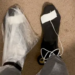 A person's feet are shown, one covered in a plastic bag, the other in a black sock with a white string and tape. These are makeshift vapor barrier liner socks for a DIY project.