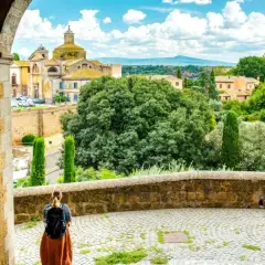 A traveler pauses, backpack ready, gazing at the historic Italian landscape. This view, perhaps on a journey from Viterbo to Rome, showcases ancient architecture and lush greenery under a bright sky.