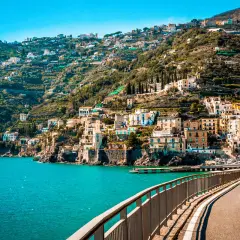 A scenic road curves along the coast, offering breathtaking views of colorful buildings clinging to a hillside. The turquoise sea sparkles below, a perfect backdrop for an unforgettable Amalfi drive.