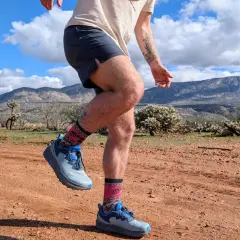 Runner's legs in Altra Timp 6 shoes and patterned socks, mid-stride on a dusty desert trail with mountains in the background.