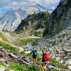Hikers ascend a rocky path alongside a stream, with majestic mountains in the distance, on the Haute Route Pyrenees.