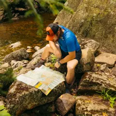 A hiker consults a map by a stream, perhaps planning his next move after some bushwhacking.