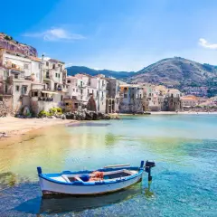 A charming fishing boat bobs in clear turquoise waters off the coast of Cefalù, a picturesque town with colorful buildings cascading down a hillside, a must-see among places to visit in Sicily.