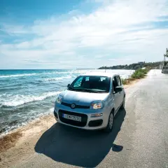A silver Fiat Panda parked on a coastal road in Greece. The turquoise sea laps the shore beside the car, suggesting this is the perfect way to explore.