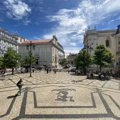 A sunny day trip to Lisbon, exploring a beautiful square with intricate mosaic paving and historic buildings under a blue, cloudy sky.