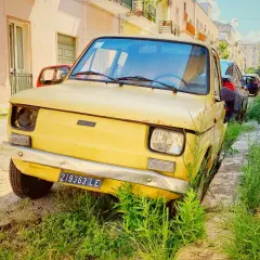 A vintage yellow Fiat, a symbol of driving in Puglia, sits parked on a sun-drenched street, overgrown with lush green grass.