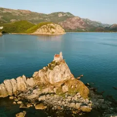 A tiny church perches on a rocky islet, a picturesque stop on a Montenegro itinerary. Mountains rise behind the tranquil blue bay.