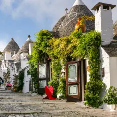 A person walks down a charming street lined with iconic trulli houses, lush greenery, and shops. This picturesque scene makes you wonder, which is the best town to stay in Puglia?