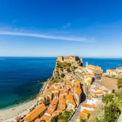A stunning view of Scilla, Calabria, with its colorful houses cascading down to a beach and the ancient Ruffo Castle perched on a cliff overlooking the turquoise sea. This is what to see in Calabria!