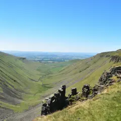 A vast green valley carved by a winding stream, with rocky outcrops in the foreground. This is a classic view along the Pennine Way, under a clear blue sky.