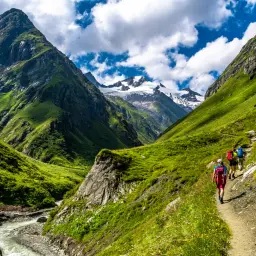 Hikers on a trail amidst lush green mountains, experiencing the beauty of long distance hikes in Europe. Snow-capped peaks loom in the distance.
