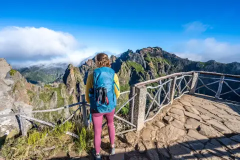 A hiker with a blue backpack enjoys a breathtaking view during a Madeira hike. Jagged, green mountains rise under a bright blue sky.