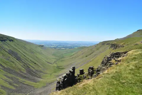 A vast green valley carved by a winding stream, with rocky outcrops in the foreground. This is a classic view along the Pennine Way, under a clear blue sky.