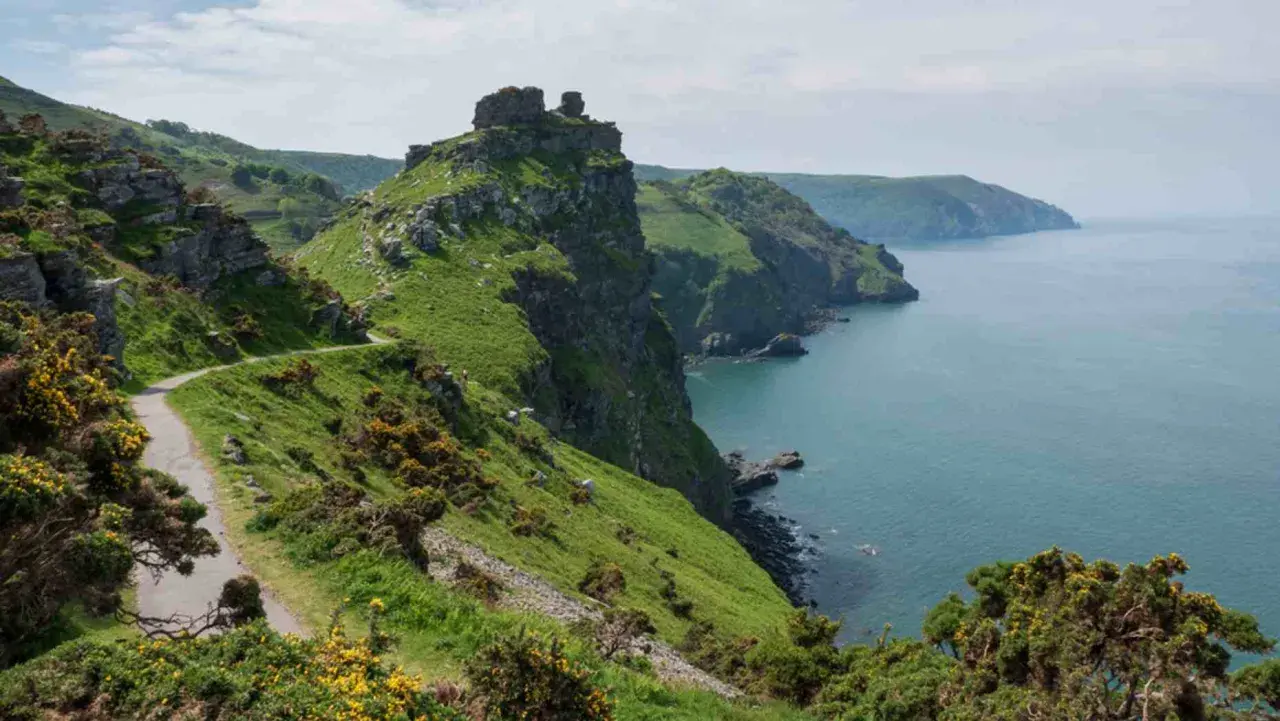 A winding path hugs a grassy cliff overlooking the sea. This is the salt path, a scenic route with rugged beauty.