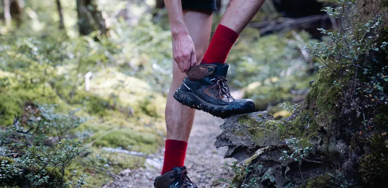 A hiker adjusts their muddy Keen shoes, showing signs of wear and tear, as they step onto a moss-covered log in a forest.