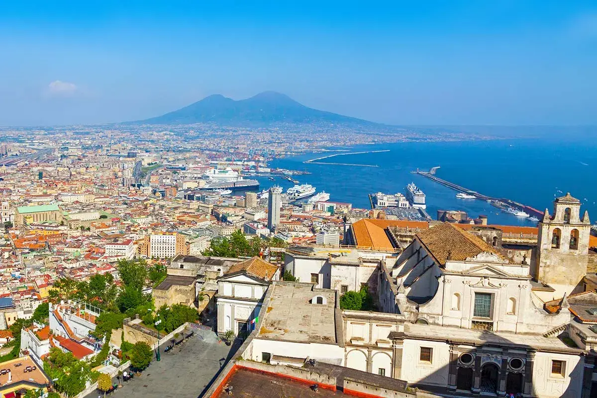 One day in Naples, a panoramic view reveals the city's sprawl, the bustling port with cruise ships, and the majestic Mount Vesuvius in the distance under a clear blue sky.