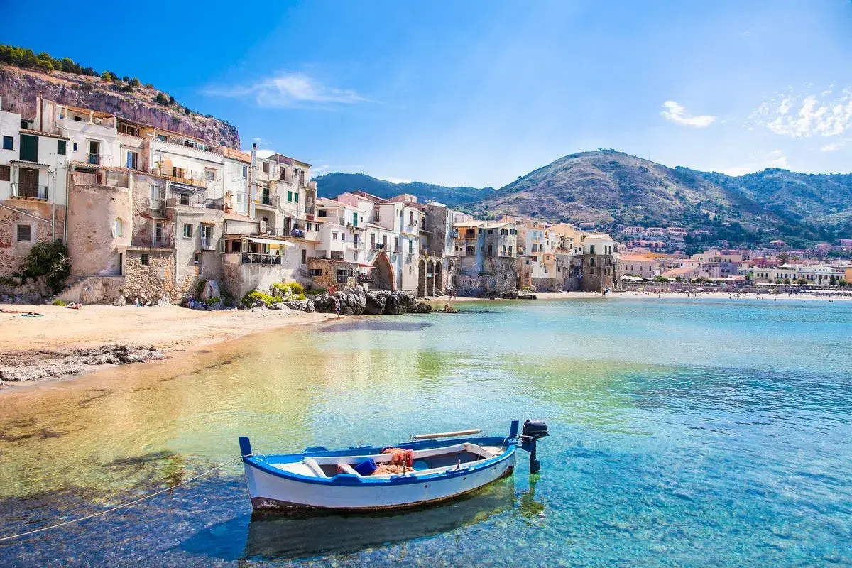 A charming fishing boat bobs in the clear turquoise water off the coast of Cefalù, a picturesque town with colorful buildings cascading down a hillside, making it one of the most beautiful places to go in Sicily.