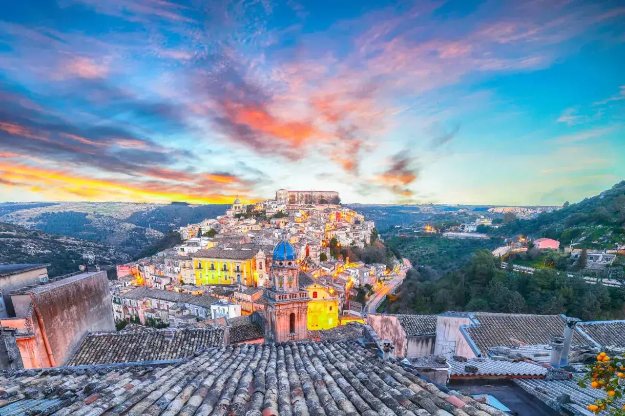 A picturesque Italian town perched on a hill, bathed in the warm glow of sunset. This view captures the essence of **two weeks in Sicily**, with its charming architecture and vibrant sky.