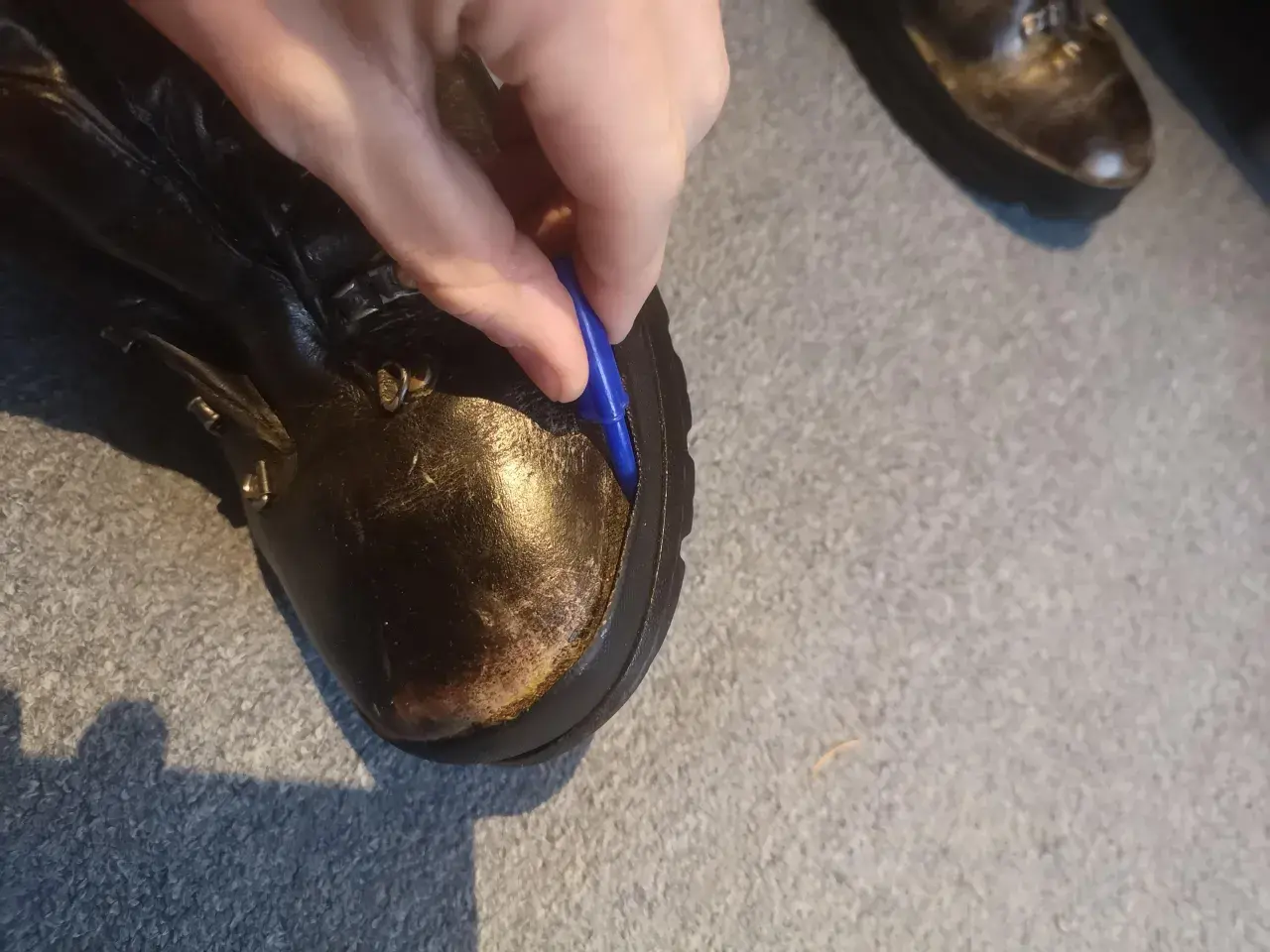 A hand uses a blue tool to pry open a seam on a leather boot, showing how to fix boots that are coming apart.