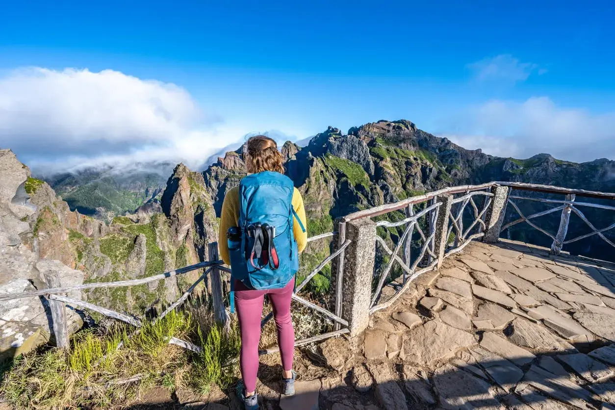 A hiker with a blue backpack enjoys a breathtaking view during a Madeira hike. Jagged, green mountains rise under a bright blue sky.