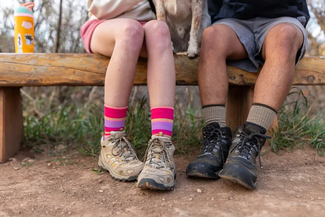 Colorful striped and neutral socks to wear with hiking boots, worn by two people sitting on a wooden bench with hiking boots on.