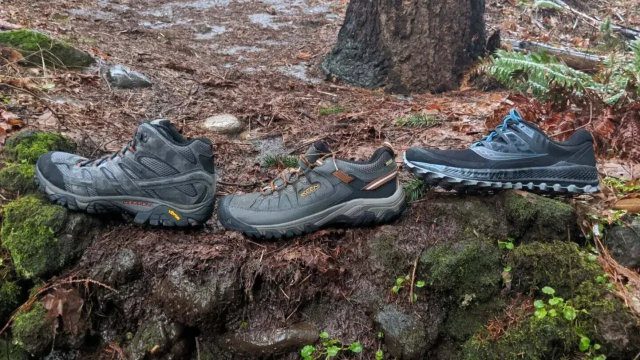 Three pairs of shoes sit on a mossy, wet forest floor: a mid-height hiking boot, a low-cut hiking shoe, and a pair of trail running shoes.