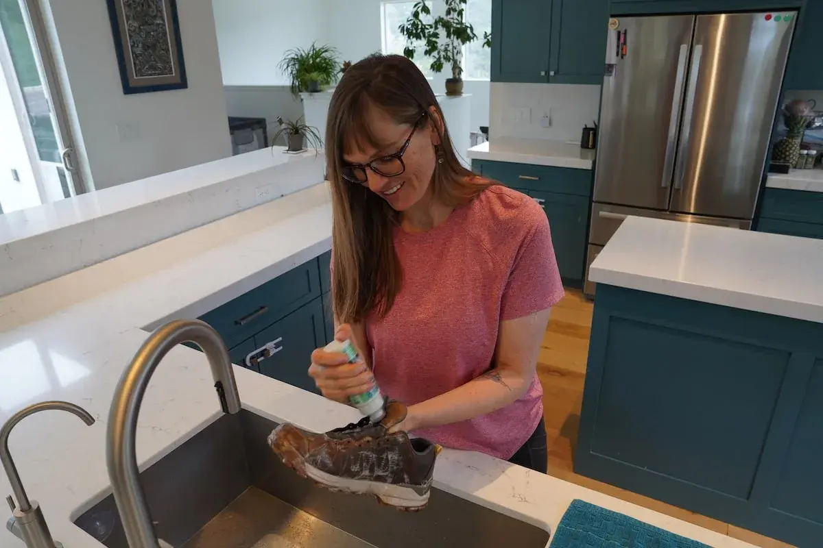 A woman cleans a dirty boot in a kitchen sink, preparing it for the washer.
