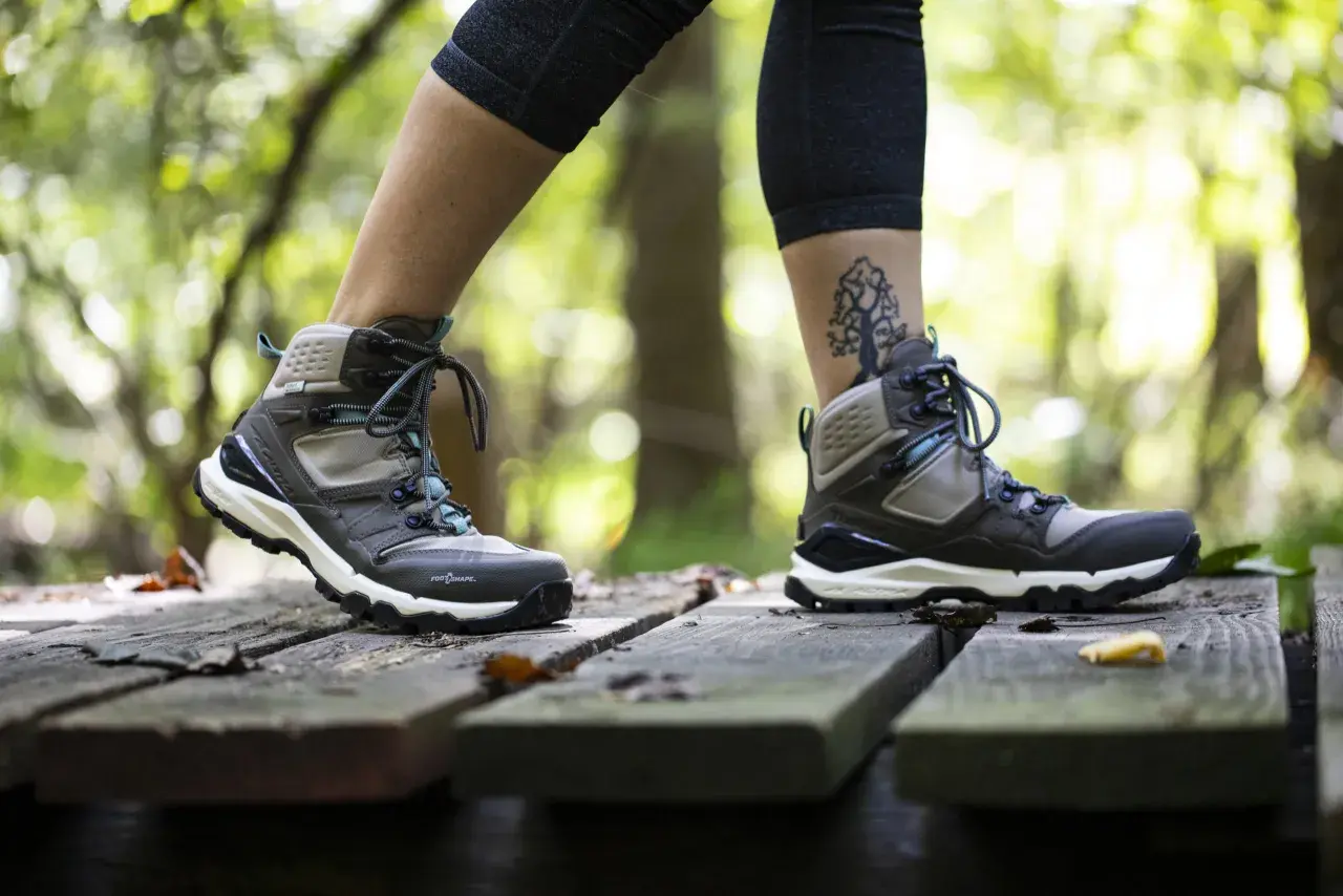 A person walks on a wooden path in the forest, wearing hiking boots. One foot half size bigger than the other, with a tree tattoo on the ankle.