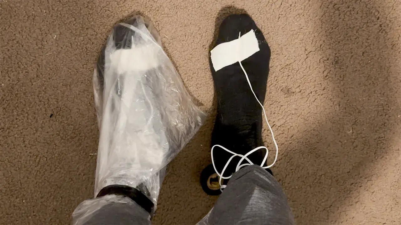 A person's feet are shown, one covered in a plastic bag, the other in a black sock with a white string and tape. These are makeshift vapor barrier liner socks for a DIY project.