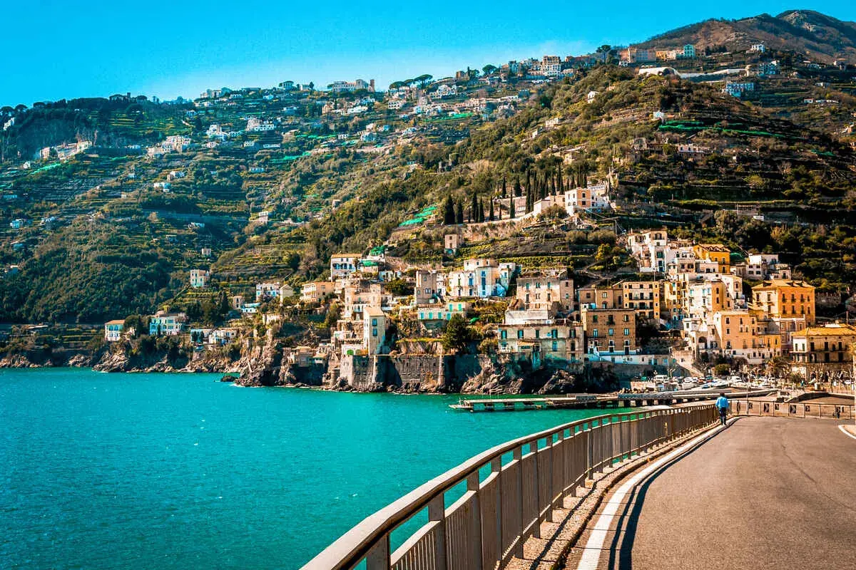 A scenic road curves along the coast, offering breathtaking views of colorful buildings clinging to a hillside. The turquoise sea sparkles below, a perfect backdrop for an unforgettable Amalfi drive.