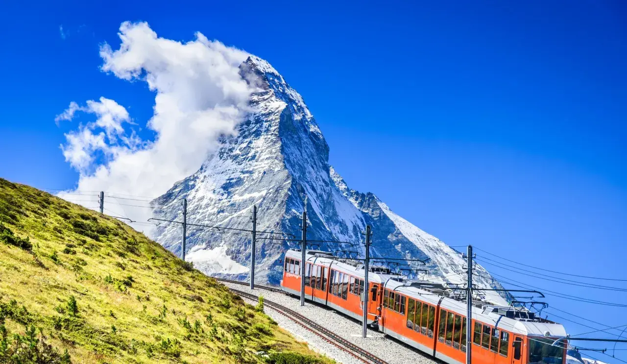 An orange cogwheel train ascends a grassy slope towards the majestic Matterhorn, a perfect scene for hiking in Switzerland.