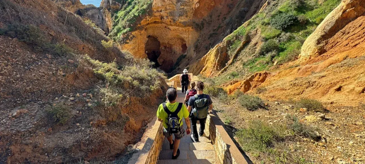 Hikers descend a stone staircase on a scenic trail in Portugal, leading towards a large cave entrance nestled within rugged, sun-drenched cliffs.