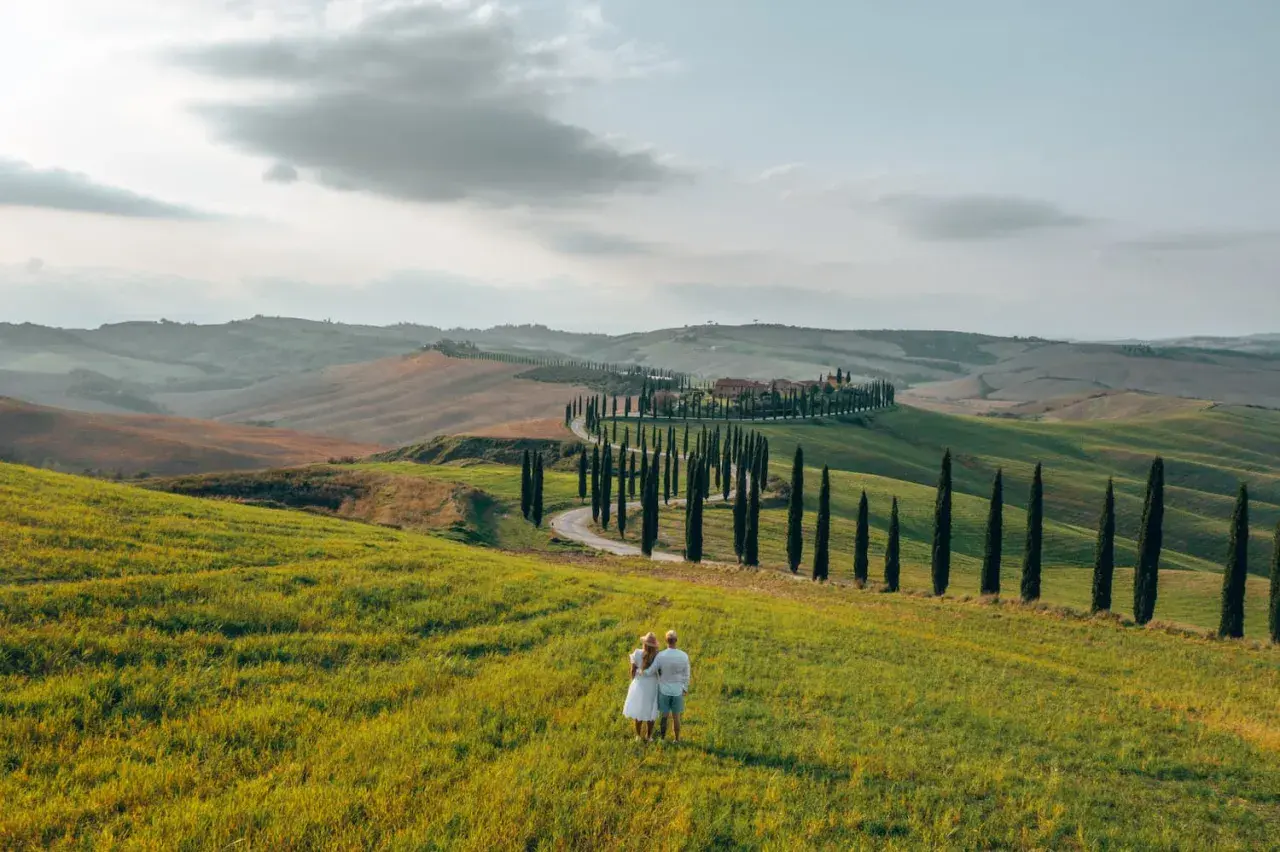 A couple enjoys the view of rolling hills and a cypress-lined road, perfect for renting a car in Tuscany.