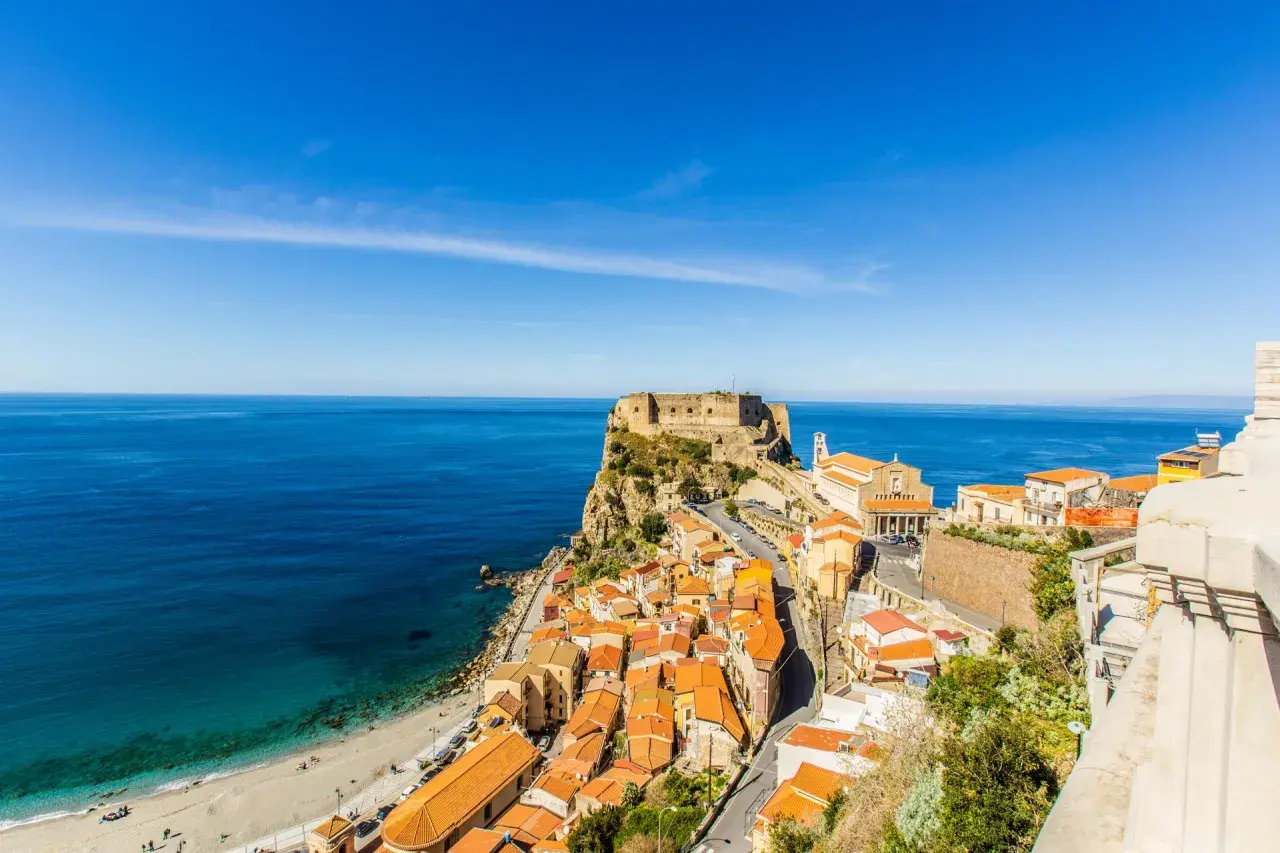 A stunning view of Scilla, Calabria, with its colorful houses cascading down to a beach and the ancient Ruffo Castle perched on a cliff overlooking the turquoise sea. This is what to see in Calabria!