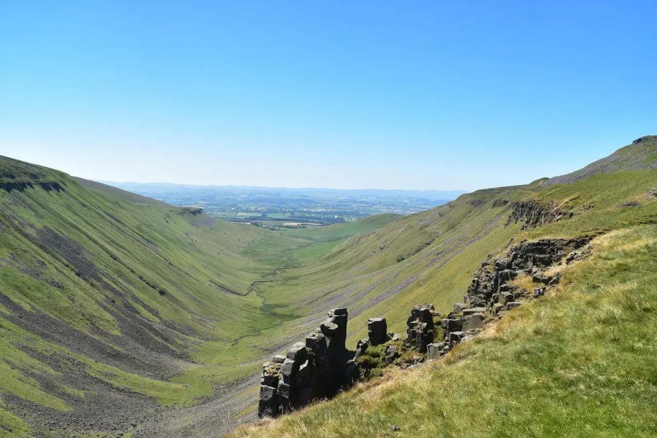A vast green valley carved by a winding stream, with rocky outcrops in the foreground. This is a classic view along the Pennine Way, under a clear blue sky.