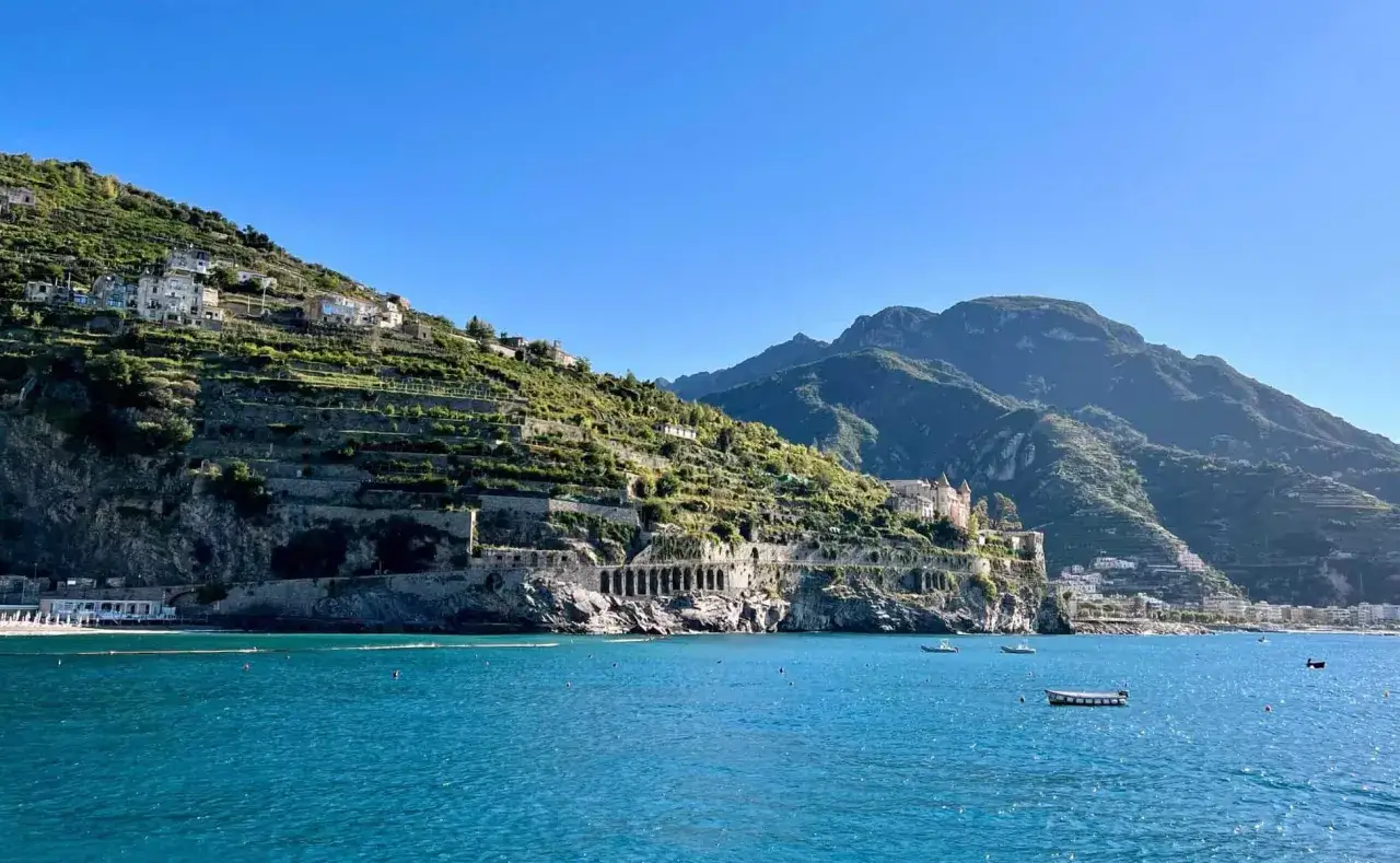 A scenic view from a Naples to Amalfi Coast ferry, showcasing terraced hillsides dotted with villas, ancient ruins, and the sparkling blue sea.